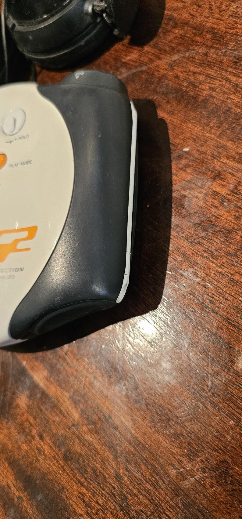 Op de afbeelding: Een draagbaar elektronisch apparaat met een mat donkergrijs zijpaneel en een wit bovenoppervlak. De bovenkant is voorzien van een ronde knop met het label HOLD, een PLAY MODE-knop en oranje tekst met het woord PROTECTION.