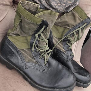 May include: A pair of black and olive green military-style combat boots with lace-up closures and a matching olive green beanie featuring a circular patch with the text United States Navy embroidered in the center of the fabric cap.