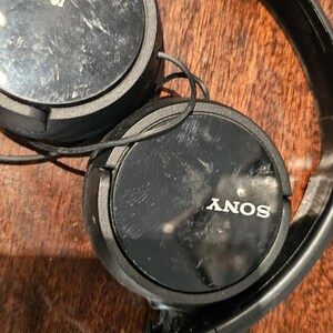Op de afbeelding: Zwarte Sony over-ear hoofdtelefoon met een matzwarte hoofdband en ronde oorschelpen met een glanzende zwarte afwerking. Het Sony-logo is in het wit gedrukt in het midden van elke oorschelp. Dunne zwarte draden verbinden de twee oorschelpen.