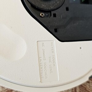 Op de afbeelding: Een wit cirkelvormig apparaat met een zwart centraal schijfmechanisme met zichtbare schroeven. Op het oppervlak is een wit rechthoekig label bevestigd met de gedrukte tekst: NEVER TOUCH THE LENS. NE JAMAIS TOUCHER LA LENTILLE.