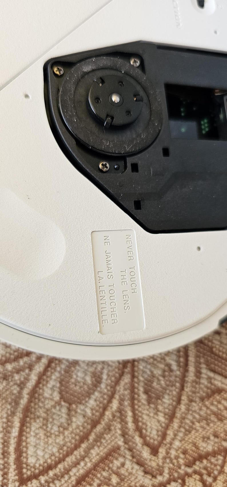 Op de afbeelding: Een wit cirkelvormig apparaat met een zwart centraal schijfmechanisme met zichtbare schroeven. Op het oppervlak is een wit rechthoekig label bevestigd met de gedrukte tekst: NEVER TOUCH THE LENS. NE JAMAIS TOUCHER LA LENTILLE.