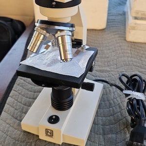 May include: A white and black microscope with silver lenses. The microscope has a black power cord and the brand name "National" is visible on the base. The lenses are labeled 10/0.25 and 40/0.65.