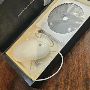 May include: An off-white Apple mouse with a cord, packaged in a black box. The box contains a CD and is labeled "Designed by Apple in California."