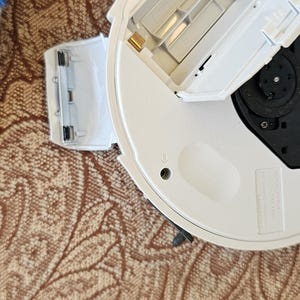 Op de afbeelding: Een wit rond elektronisch apparaat met een open batterijcompartiment dat gouden metalen contactpunten toont. Het apparaat heeft een zwart centraal onderdeel en gegoten tekst met de tekst NEVER TOUCH THE LENS. NE JAMAIS TOUCHER LA LENTILLE.