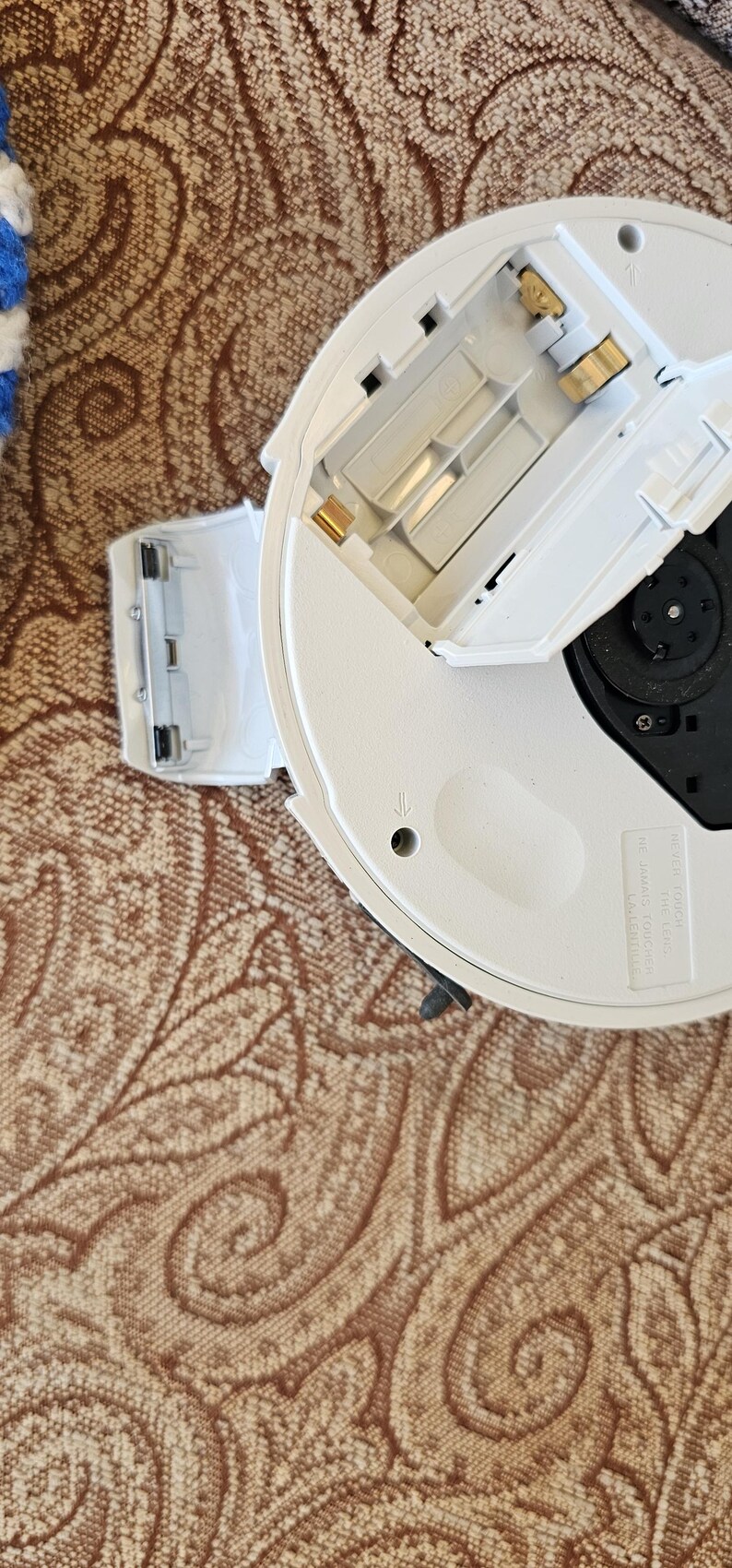 Op de afbeelding: Een wit rond elektronisch apparaat met een open batterijcompartiment dat gouden metalen contactpunten toont. Het apparaat heeft een zwart centraal onderdeel en gegoten tekst met de tekst NEVER TOUCH THE LENS. NE JAMAIS TOUCHER LA LENTILLE.