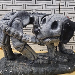 May include: A gray concrete statue of a dog and a person. The dog is on its hind legs and the person is leaning down to pet the dog. The statue is on a gray base.
