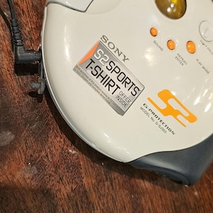 Op de afbeelding: Een ronde, gebroken witte Sony S2 Sports cd-speler, model D-SJ303, met een geel rond venster, oranje bedieningsknoppen en een zwart hoofdtelefoonsnoer. Het apparaat heeft een G-Protection-label en een promotionele sticker voor een T-shirt.