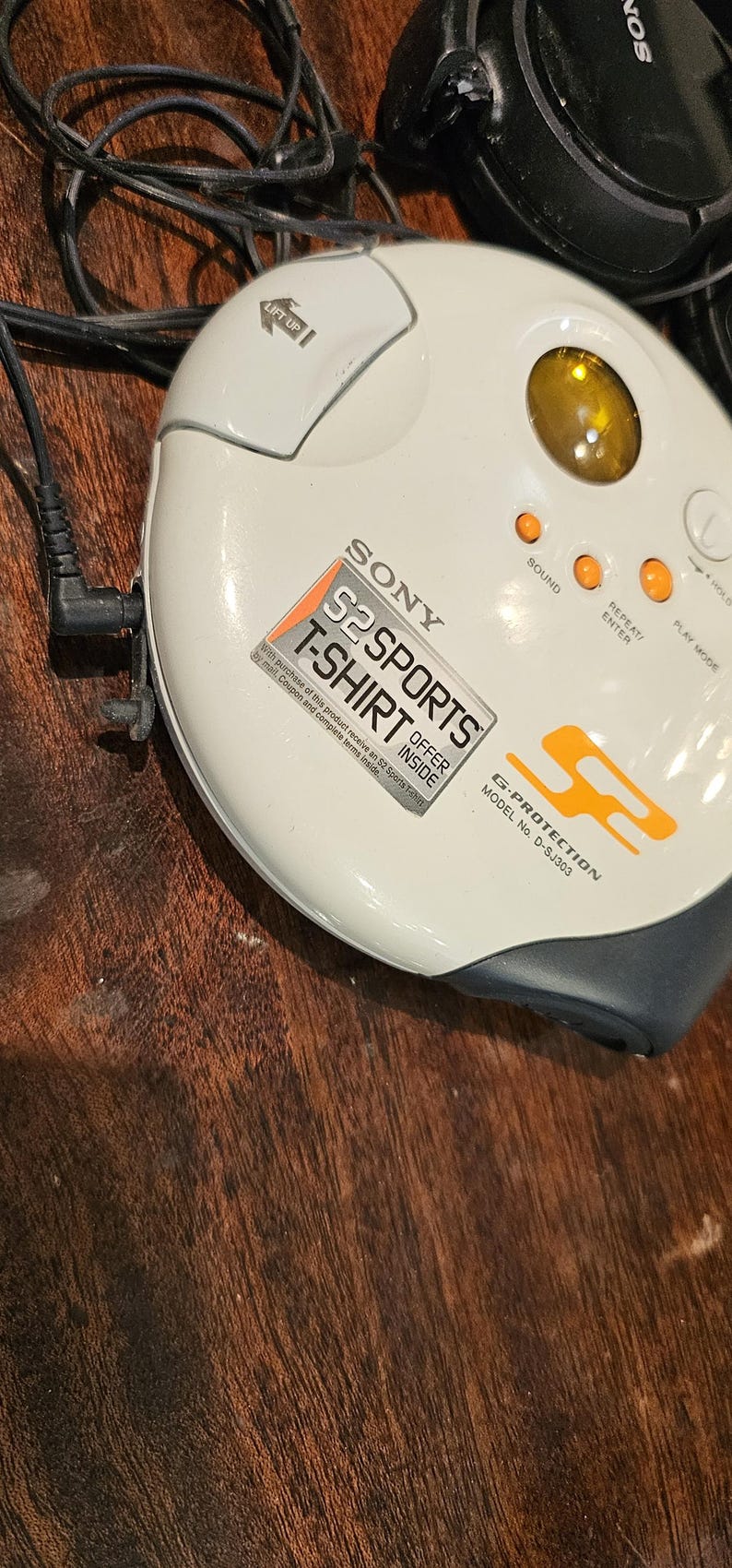 Op de afbeelding: Een ronde, gebroken witte Sony S2 Sports cd-speler, model D-SJ303, met een geel rond venster, oranje bedieningsknoppen en een zwart hoofdtelefoonsnoer. Het apparaat heeft een G-Protection-label en een promotionele sticker voor een T-shirt.