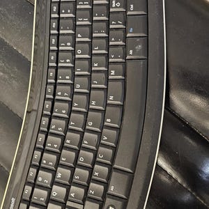May include: A black Microsoft ergonomic keyboard with white trim. The keyboard has a curved design and features standard QWERTY keys, function keys, and a Windows key. The keyboard is placed on a black surface.
