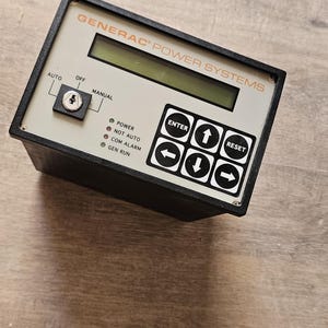 May include: A rectangular Generac Power Systems control panel with a black casing and a light gray faceplate. It features a digital display, a key-operated switch for Auto, Off, and Manual modes, four status indicator lights, and a keypad with Enter, Reset, and directional arrow buttons.