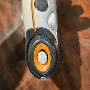 Op de afbeelding: Een wit en donkergrijs draagbaar elektronisch apparaat met een oranje accentlijn, een zwarte volumeknop met het label VOL en een ronde bedieningsinterface met een oranje ring en een centrale knop met een afspeelpictogram.