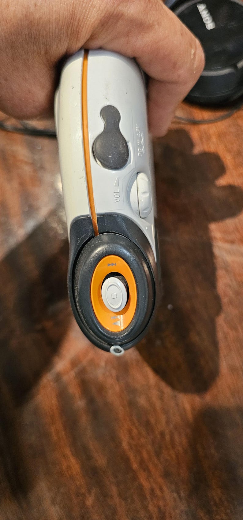 Op de afbeelding: Een wit en donkergrijs draagbaar elektronisch apparaat met een oranje accentlijn, een zwarte volumeknop met het label VOL en een ronde bedieningsinterface met een oranje ring en een centrale knop met een afspeelpictogram.