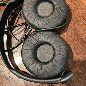 Op de afbeelding: Zwarte bedrade hoofdtelefoon met twee ronde, getextureerde oorkussens met een matte afwerking. De hoofdband is gemaakt van zwart plastic en bevat een dunne, ge&iuml;ntegreerde zwarte audiokabel die de twee oorschelpen met elkaar verbindt.