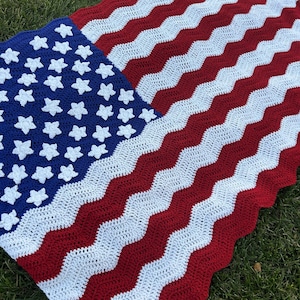 May include: A handmade crochet blanket in the style of the American flag. The blanket features a blue square with white star appliques in the upper left corner, and red and white wavy stripes across the rest of the blanket.