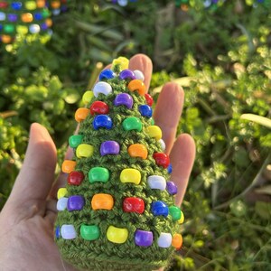 May include: A small, handmade green crochet Christmas tree decorated with colorful beads. The tree is adorned with beads in various colors, including red, orange, yellow, purple, blue, and white. The tree is held in a hand against a background of green foliage.