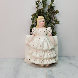 May include: A white ceramic wall planter shaped like a girl in a white dress with red and green floral print. The girl has blonde hair and is holding a bouquet of green leaves.