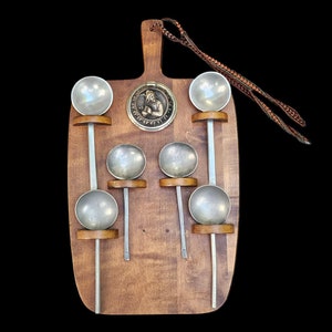 May include: A wooden board with six silver ladles hanging from it. The ladles have wooden handles and are attached to the board with metal hooks. The board has a decorative metal medallion in the center.