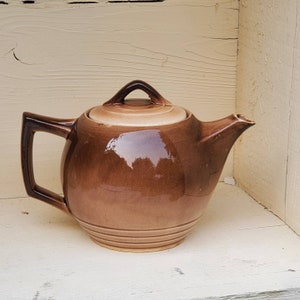 May include: A brown ceramic teapot with a lid and a rounded body. The teapot has a light brown glaze and a darker brown glaze on the bottom.