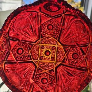 May include: A vibrant red, round glass ornament with intricate geometric and floral patterns. The ornament features a detailed, cut-glass design with a central starburst and radiating shapes. The glass has a glossy finish and is suspended by a small loop.
