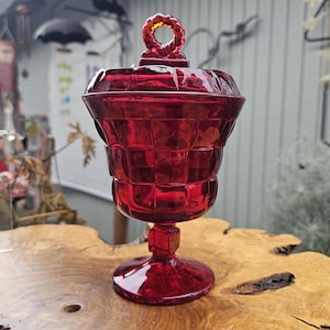 May include: A red glass candy dish with a lid and a ring handle. The dish has a pedestal base and a square pattern.