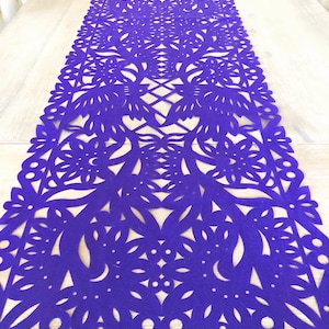 May include: Purple table runner with a delicate, intricate cut-out design featuring flowers and leaves. The runner is made of a thick, felt-like material.