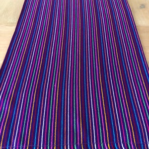 May include: A purple table runner with colorful stripes and a fringe border. The stripes are in various colors, including pink, blue, green, yellow, and orange.