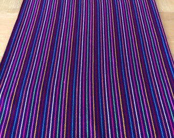 May include: A purple table runner with colorful stripes and a fringe border. The stripes are in various colors, including pink, blue, green, yellow, and orange.