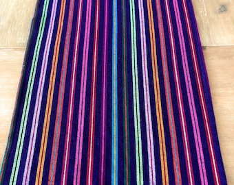 May include: A colorful table runner with a purple background and stripes of pink, red, orange, yellow, green, and blue. The runner has a fringe border.