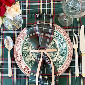 May include: A festive table setting with a green, red, and white plaid tablecloth. A folded plaid napkin is tied with a beige ribbon, placed on a floral patterned plate. Silverware and a wine glass complete the elegant arrangement.
