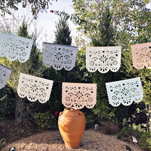 May include: A string of colorful paper cutouts, known as papel picado, hangs between trees in a garden setting. The cutouts are in various shades of pink, blue, and white, and feature intricate floral designs. A terracotta pot sits in the center of the image.