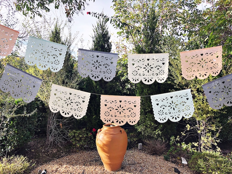 May include: A string of colorful paper cutouts, known as papel picado, hangs between trees in a garden setting. The cutouts are in various shades of pink, blue, and white, and feature intricate floral designs. A terracotta pot sits in the center of the image.
