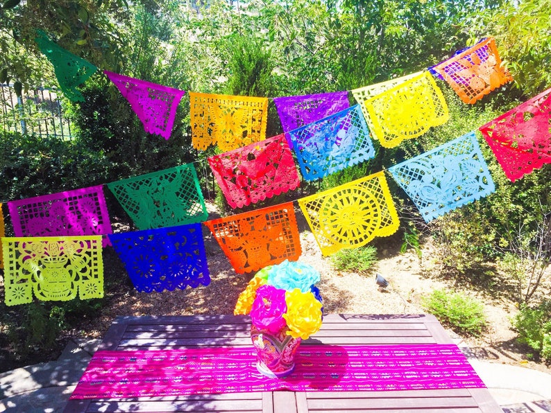 K&ouml;nnte beinhalten: Ein farbenfrohes Papierbanner mit ausgeschnittenen Designs h&auml;ngt zwischen B&auml;umen in einem Garten. Das Banner besteht aus leuchtenden Farben wie Gelb, Orange, Rot, Rosa, Lila, Blau und Gr&uuml;n. Das Banner ist zwischen B&auml;umen gespannt und h&auml;ngt &uuml;ber einem Holztisch mit einer rosa Tischdecke.