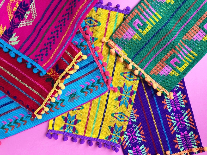 May include: Five colorful woven placemats with fringe and pom-pom trim. The placemats are in shades of pink, blue, yellow, green, and purple.