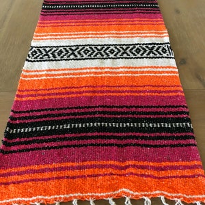 May include: A colorful Mexican table runner with stripes of orange, pink, black, and white. The runner has a fringe on the bottom edge.