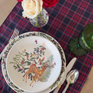 May include: A festive table setting with a red and navy plaid runner. A plate with a Christmas tree and reindeer design is set with silverware. A green glass and a vase of red and white roses complete the holiday scene.
