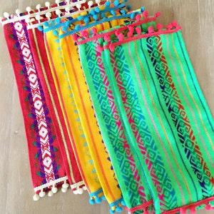 May include: Set of five colorful woven placemats with fringe and pom-pom trim. The placemats feature geometric patterns in shades of red, yellow, green, and blue.