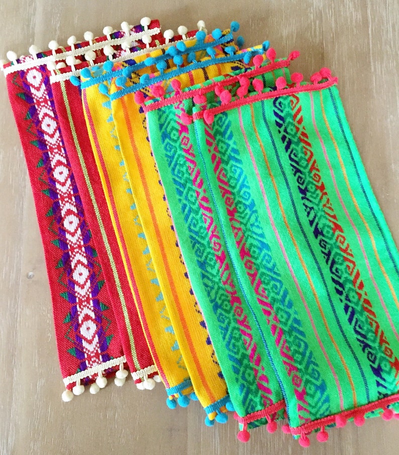 May include: Set of five colorful woven placemats with fringe and pom-pom trim. The placemats feature geometric patterns in shades of red, yellow, green, and blue.