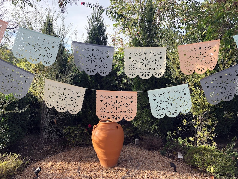 May include: A string of colorful paper banners with intricate cut-out designs hangs between trees. The banners are in shades of blue, white, and pink. A terracotta pot sits in the foreground.