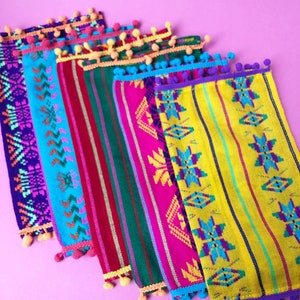 May include: A set of six colorful woven fabric placemats with fringe and pom-poms. The placemats feature geometric patterns in shades of pink, blue, green, yellow, and purple.