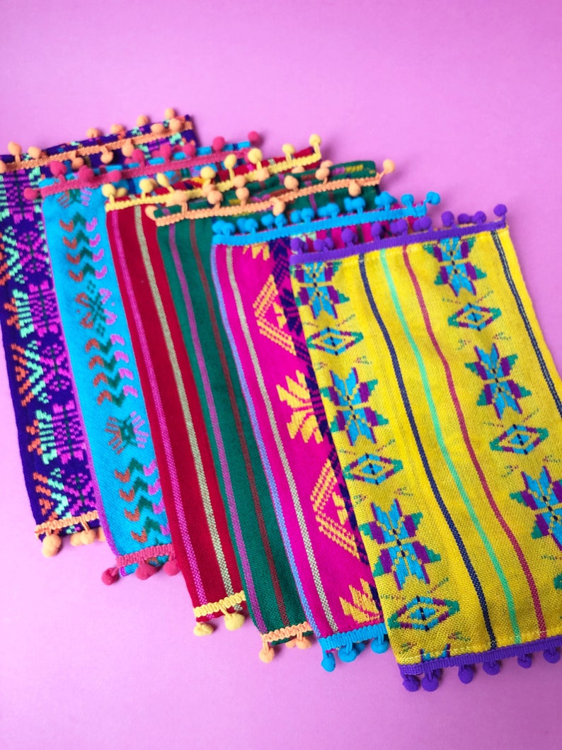 May include: A set of six colorful woven fabric placemats with fringe and pom-poms. The placemats feature geometric patterns in shades of pink, blue, green, yellow, and purple.
