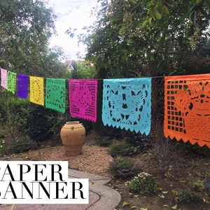 K&ouml;nnte beinhalten: Ein farbenfrohes Papierbanner mit ausgeschnittenen Designs in verschiedenen Farben, darunter Rot, Orange, Gelb, Gr&uuml;n, Blau, Pink und Lila. Das Banner h&auml;ngt im Freien vor dem Hintergrund gr&uuml;ner B&auml;ume und eines braunen Topfes.