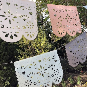May include: A string of four paper banners with cut-out designs. The banners are white, light blue, light pink, and gray. The banners are hanging from a string against a backdrop of green foliage.
