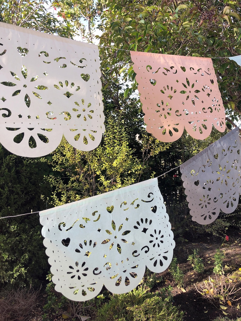 May include: A string of four paper banners with cut-out designs. The banners are white, light blue, light pink, and gray. The banners are hanging from a string against a backdrop of green foliage.