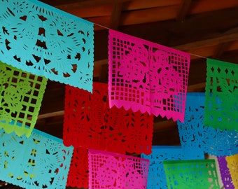 May include: A colorful string of paper decorations, known as "papel picado", hangs from a wooden ceiling. The decorations are cut in intricate patterns and feature a variety of colors, including blue, green, red, pink, and purple.