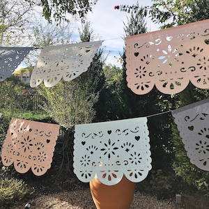 May include: A string of pastel colored paper banners with intricate cut-out designs. The banners are strung together with white string and are hanging from a tree branch. The banners are in the colors of light blue, light pink, and gray.