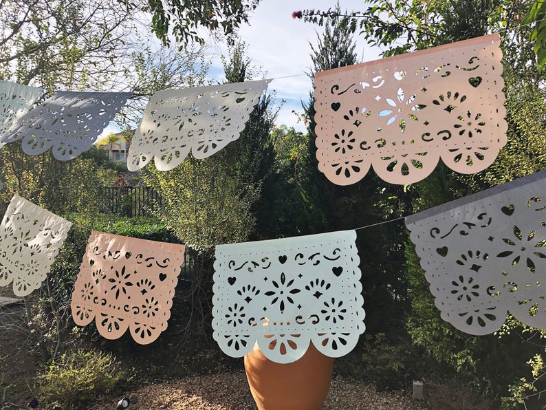 May include: A string of pastel colored paper banners with intricate cut-out designs. The banners are strung together with white string and are hanging from a tree branch. The banners are in the colors of light blue, light pink, and gray.