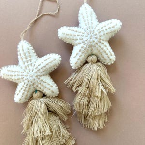 May include: Two white starfish ornaments with beige embroidered details and long beige tassels. Each starfish has a beige string for hanging. The ornaments are on a light brown background.