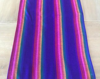 May include: A vibrant, rectangular textile with vertical stripes in shades of purple, pink, orange, yellow, and green. The textile has a deep blue background and a fringed edge with a rainbow of colors. The item appears to be a table runner or decorative scarf.