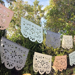 May include: A string of colorful paper banners with intricate cut-out designs, hanging outdoors against a backdrop of green trees. The banners are in shades of pink, light blue, gray, and white.