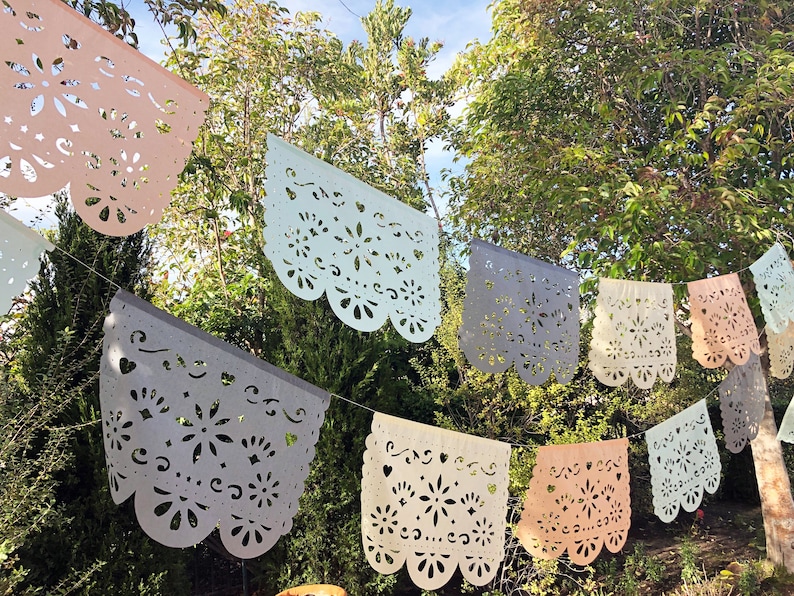 May include: A string of colorful paper banners with intricate cut-out designs, hanging outdoors against a backdrop of green trees. The banners are in shades of pink, light blue, gray, and white.